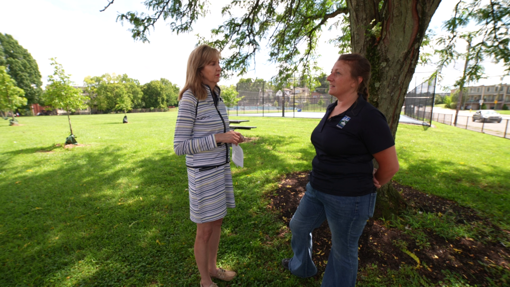 A middle aged woman with shoulder length light brown hair wearing a striped suit talks to a younger woman with braided brown hair wearing a blue polo shirt and jeans.